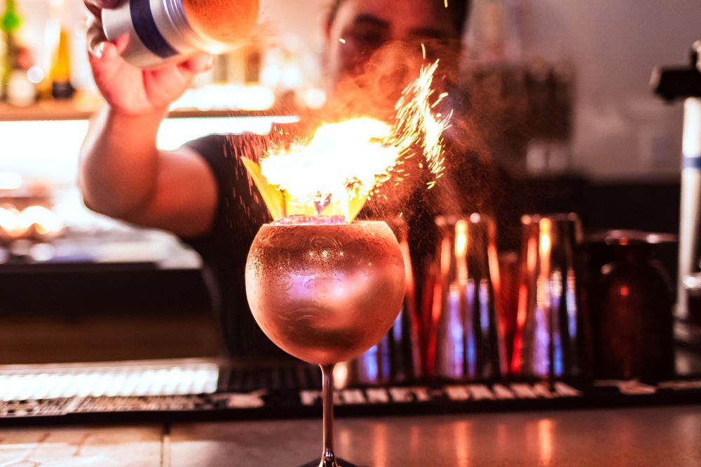 Dramatic flaming cocktail being prepared on a dark bar counter for restaurant branding photography.