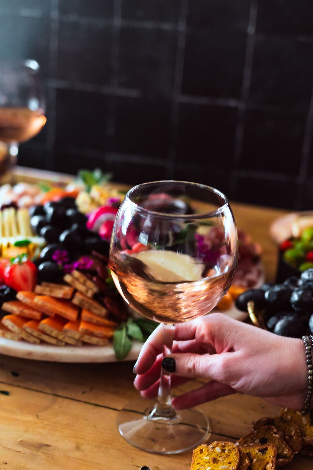 A hand holding a glass of rosé wine next to a charcuterie board with grapes and assorted meats for winery and catering branding photography.