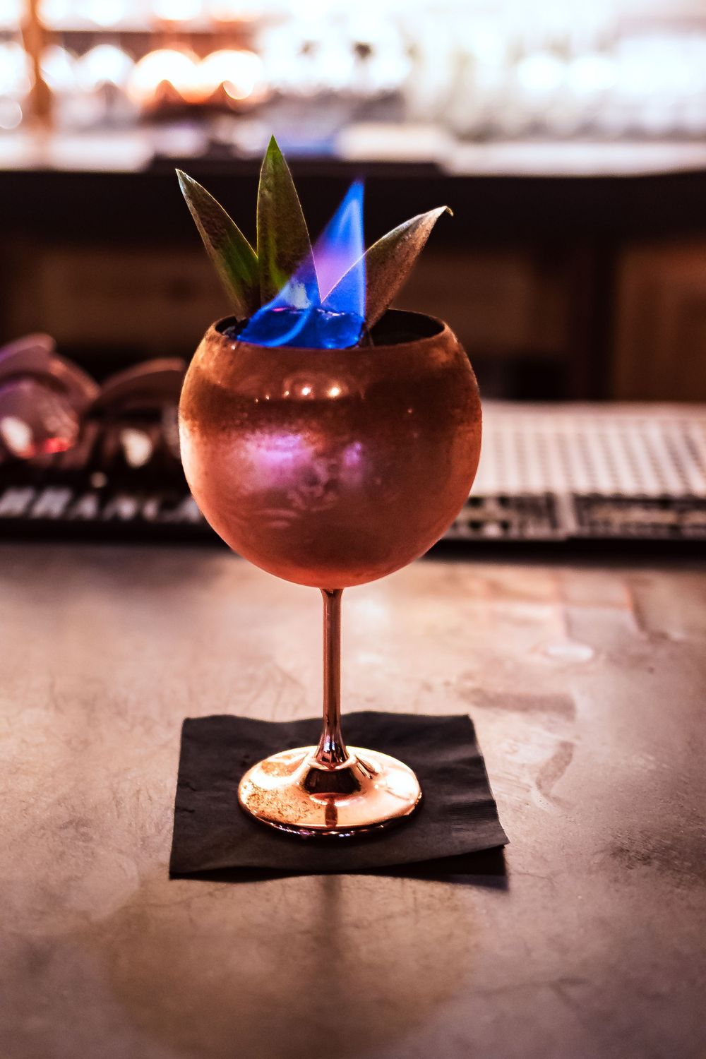 Copper cocktail glass with blue flames and leaf garnish on black napkin for restaurant branding photography.