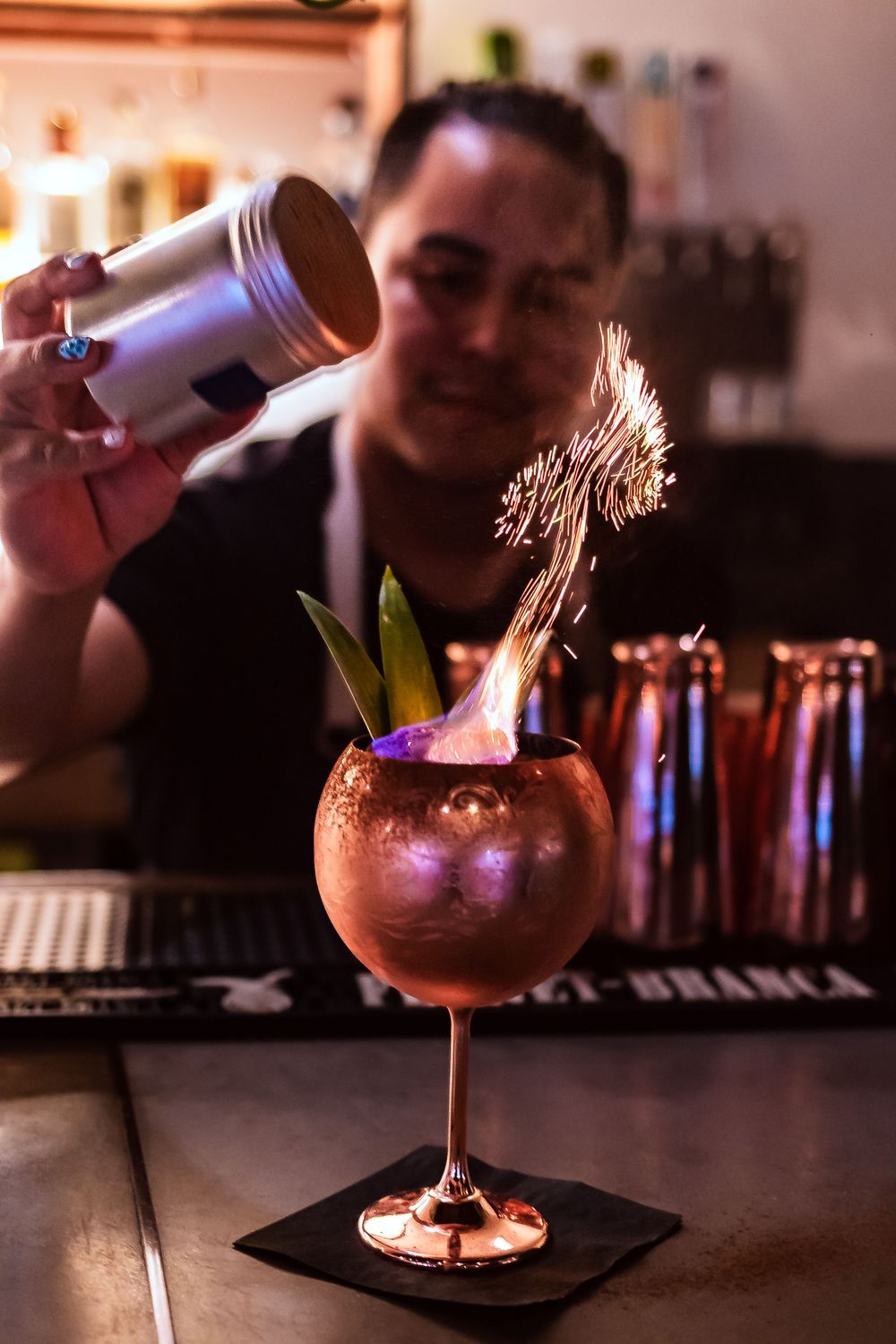 Flaming copper cocktail glass garnished with leaves on a dark bar counter for restaurant branding photography.