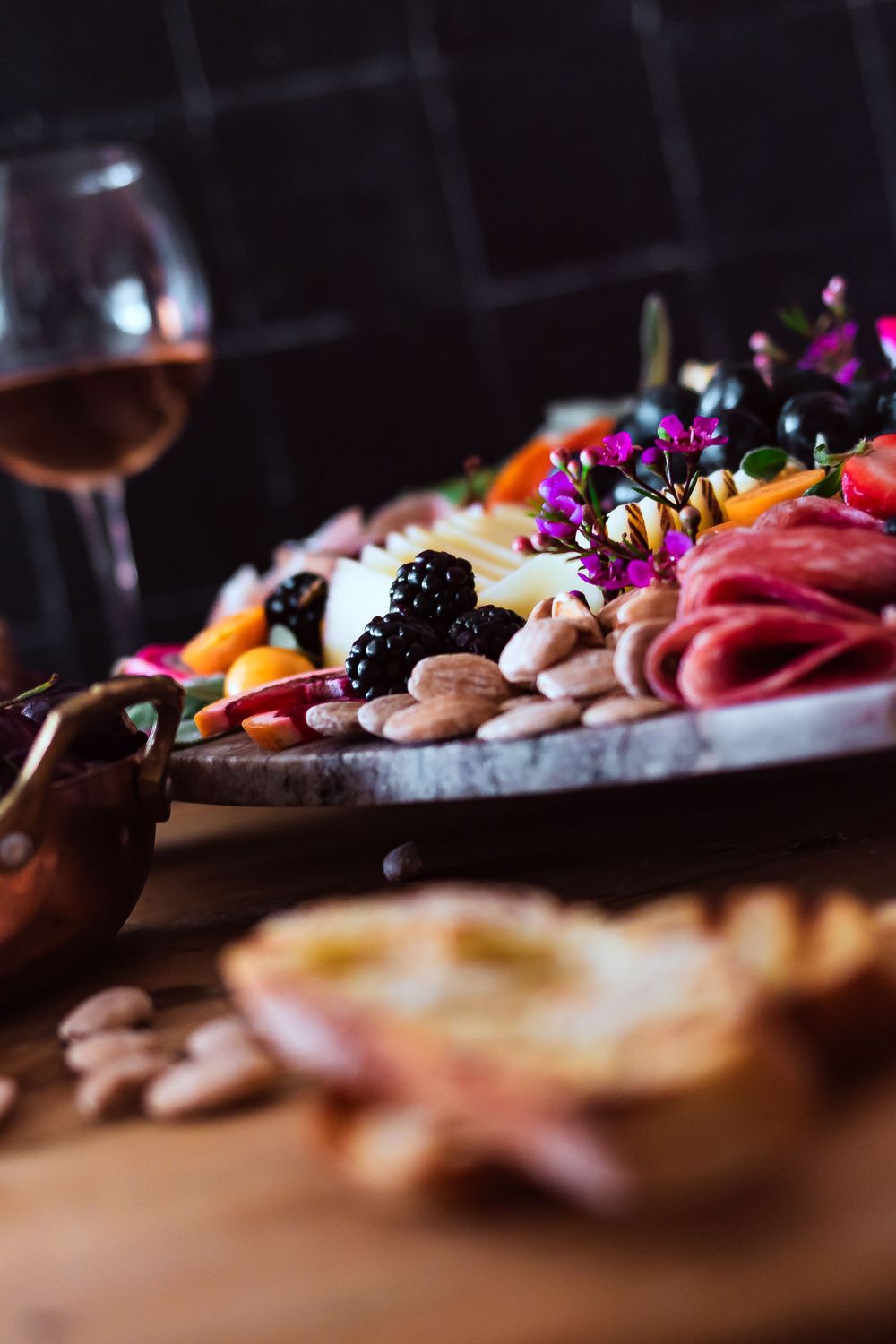Elegant charcuterie platter with wine glass, meats, fruits and edible flowers in moody lighting for winery and catering branding photography