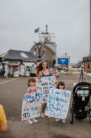 People holding handmade welcome signs near a large military ship at a naval base.