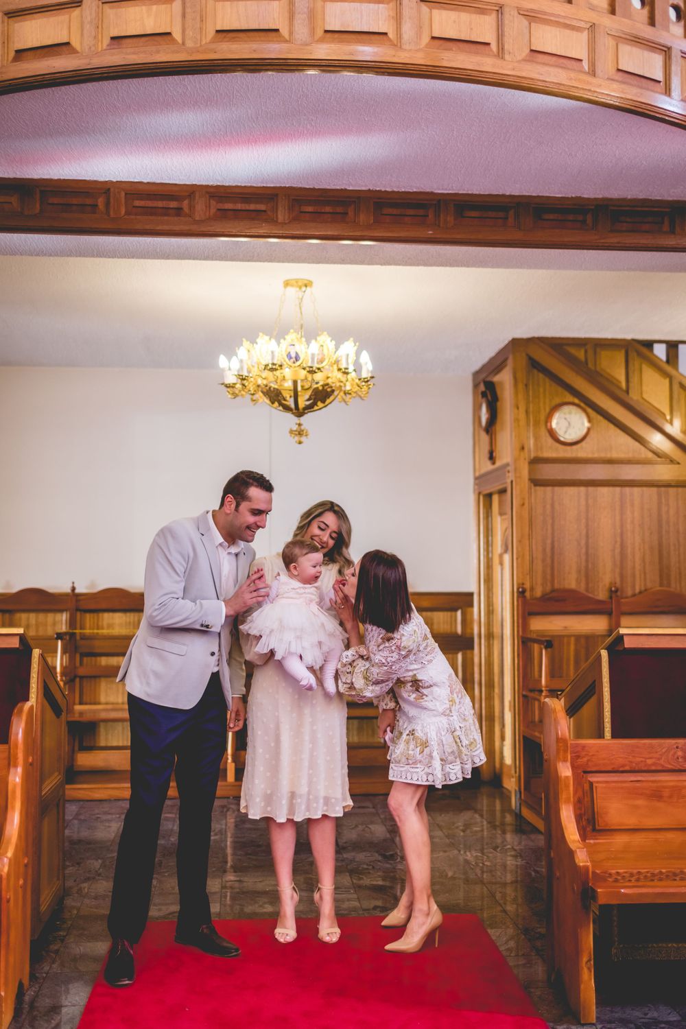 Family gathered inside a traditional church with wooden pews and ornate chandelier during a religious ceremony.