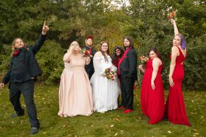 Wedding party in red and blush dresses poses joyfully outdoors with raised arms.