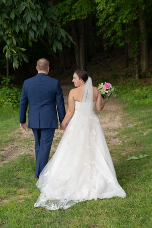 A bride in a white lace gown and her partner in a navy suit walk along a wooded path during their wedding day.