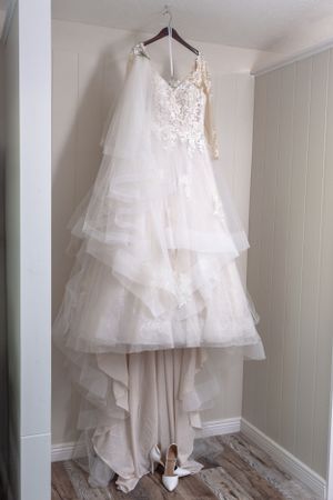 Elegant white wedding dress with lace details hangs on display against light colored wall.