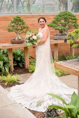 Bride in elegant lace wedding dress with long train poses by potted bonsai trees in a garden setting.