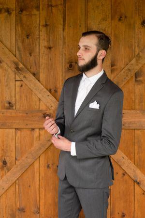 Groom in a grey suit and white pocket square poses against rustic wooden barn doors.