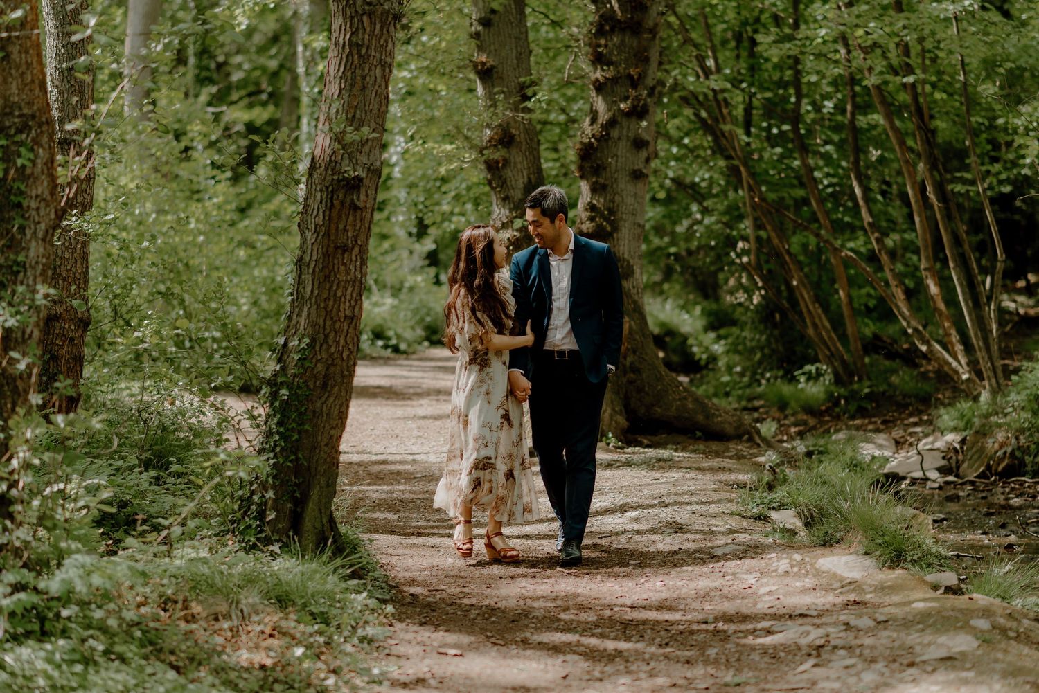 Couple strolls together along tree-lined forest path during spring engagement session.