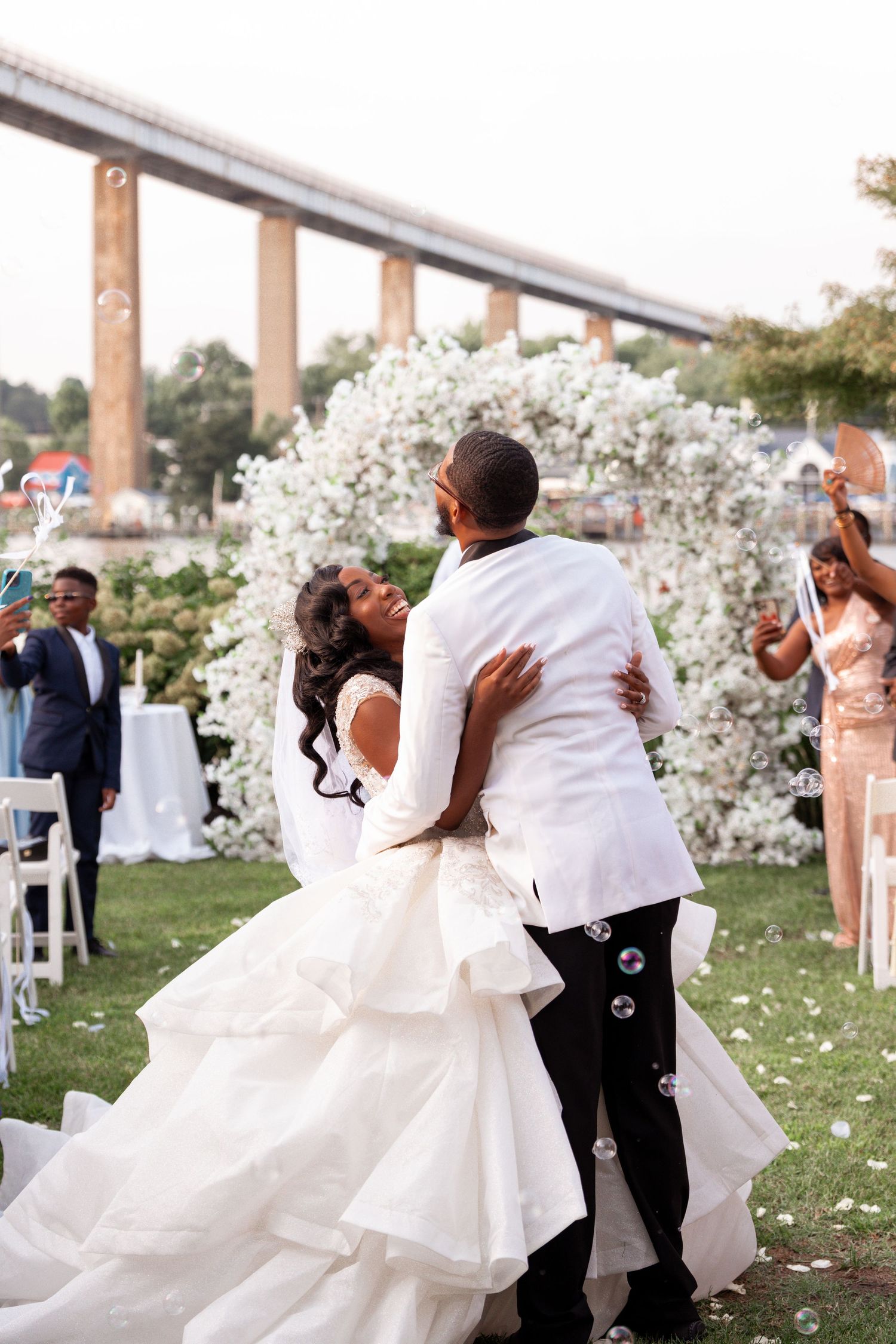 Joyful newlyweds embrace during their outdoor wedding ceremony surrounded by bubbles and a white floral arch.