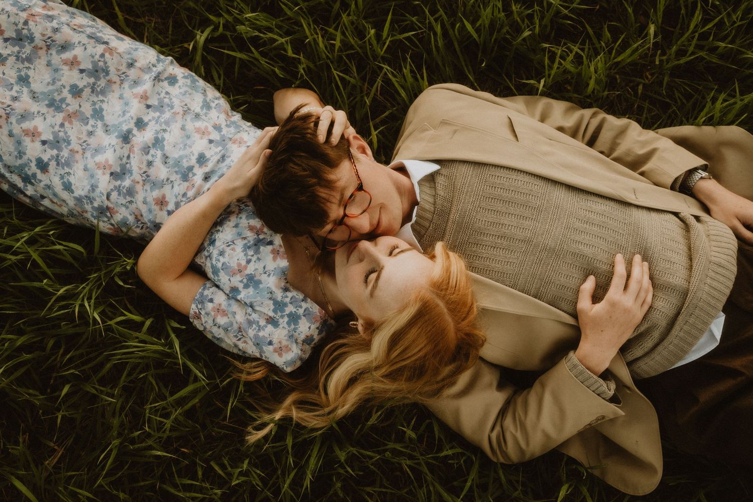Couple lying in grass wearing casual autumn attire during intimate moment.