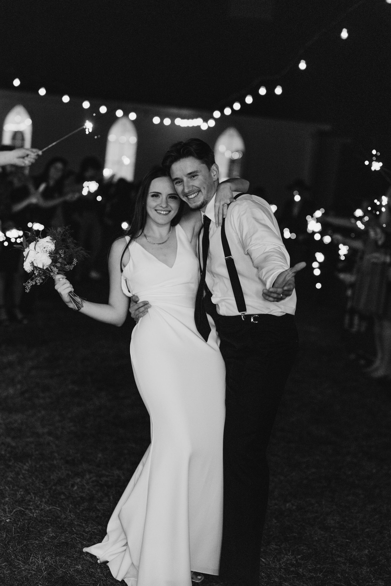Black and white photo of smiling newlyweds under string lights at night reception.
