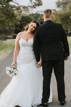 Bride in lace gown and groom in tuxedo walking together on outdoor path.