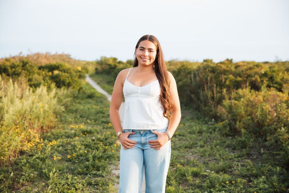 Young woman smiling in white tank and jeans on a scenic green trail path. Middletown Rhode Island. Senior photoshoot. Third beach