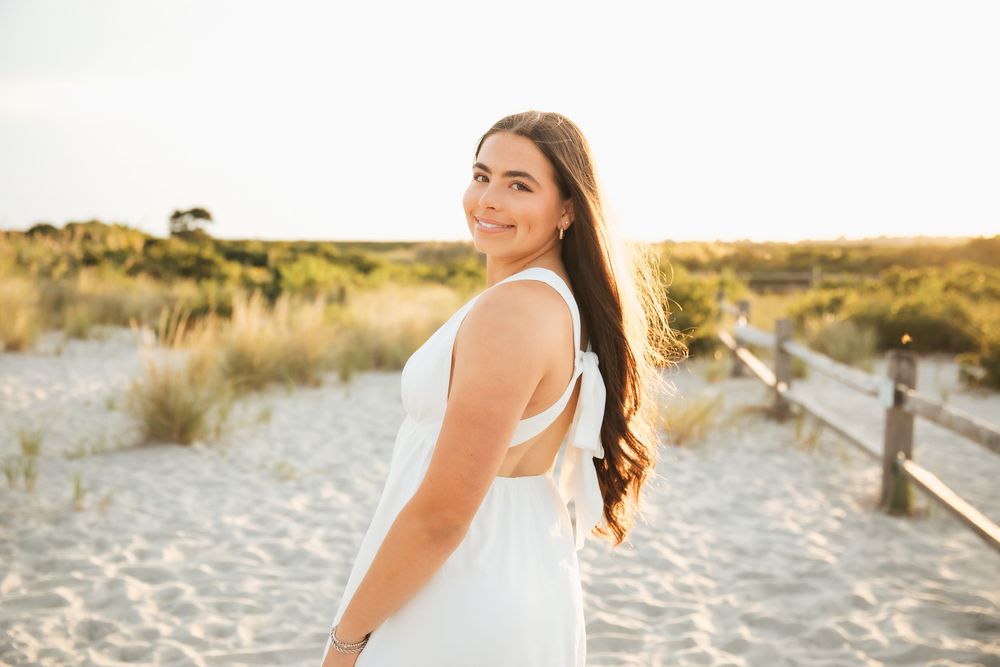 Young woman in white dress smiling on a sandy beach pathway at golden hour sunset. Middletown Rhode Island. Senior photoshoot. Third beach