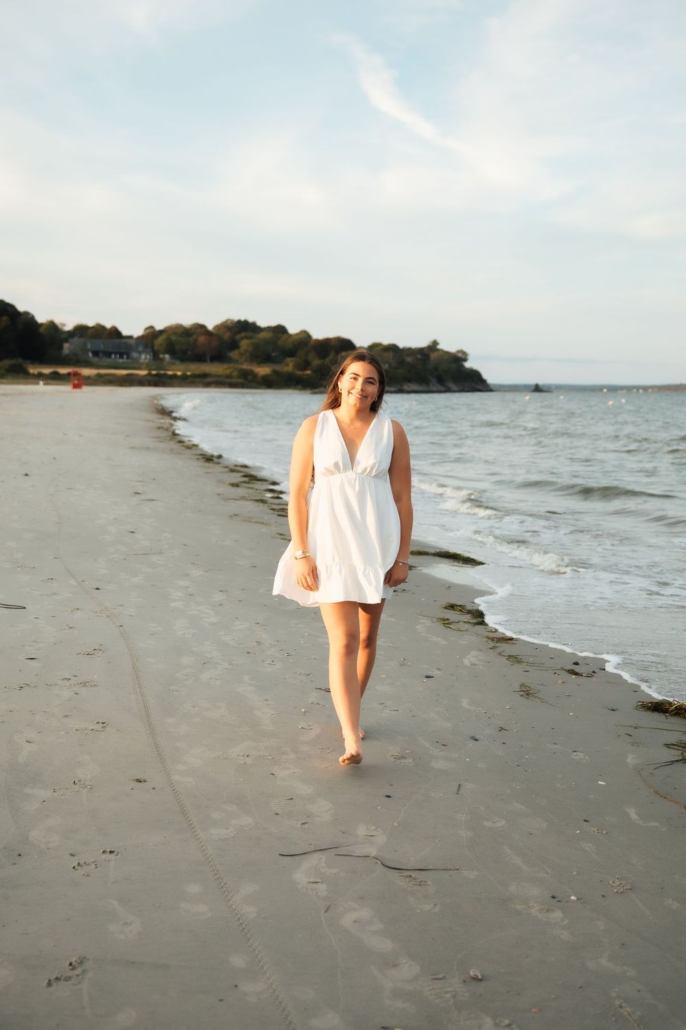 A young woman in a white sundress walks barefoot along a sandy beach. Middletown Rhode Island. Senior photoshoot. Third beach