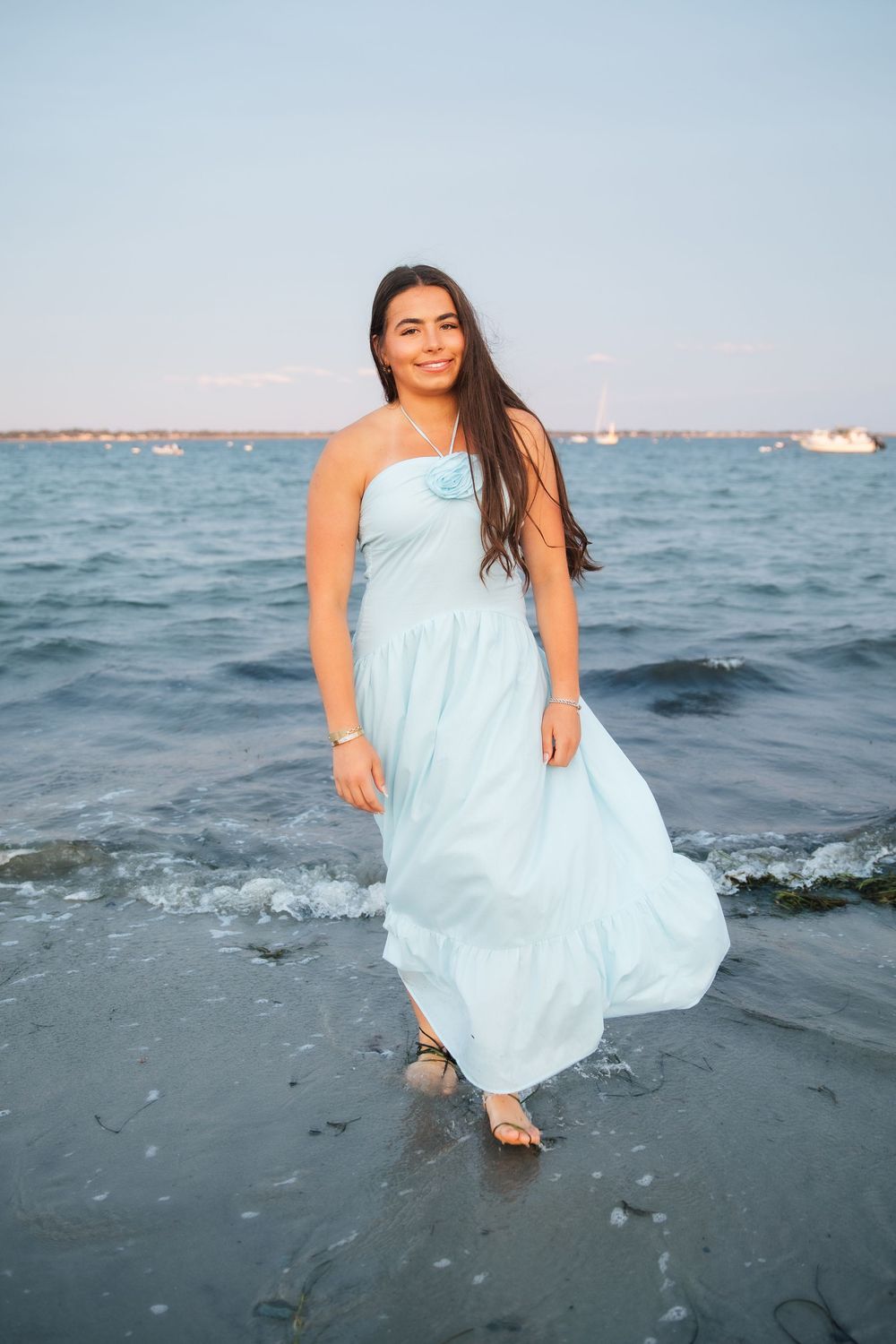 Young woman in light blue maxi dress standing in ocean waves at sunset on a serene beach. Middletown Rhode Island. Senior photoshoot
