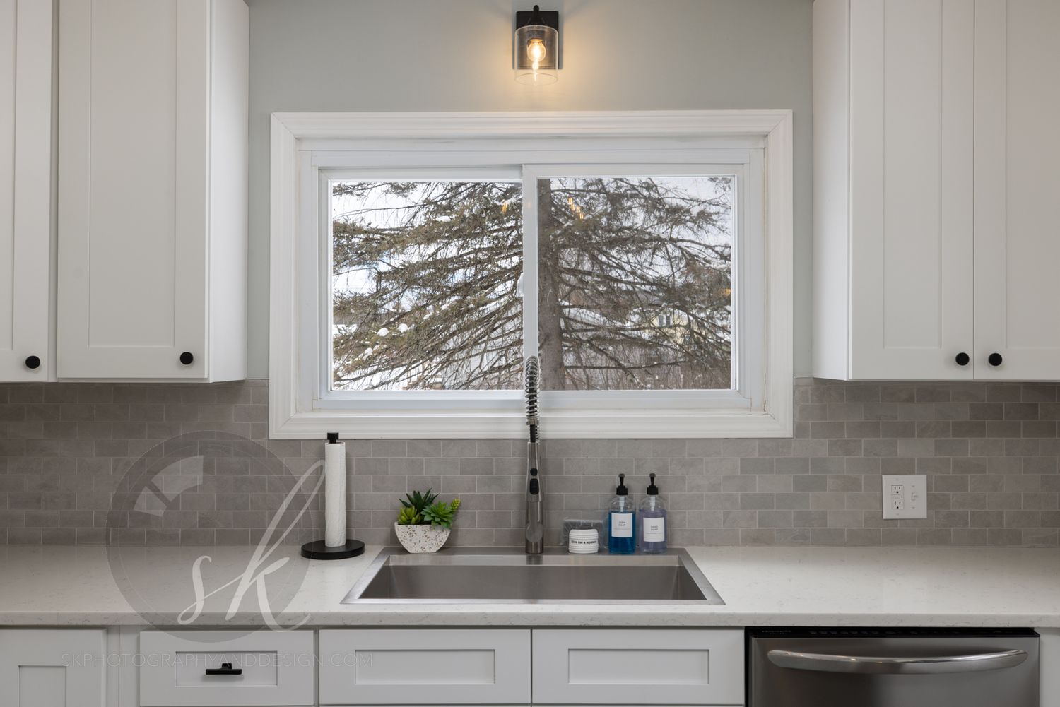 Modern kitchen window with stainless steel sink overlooks snowy evergreen trees through white-framed sliding window. Professionally taken.