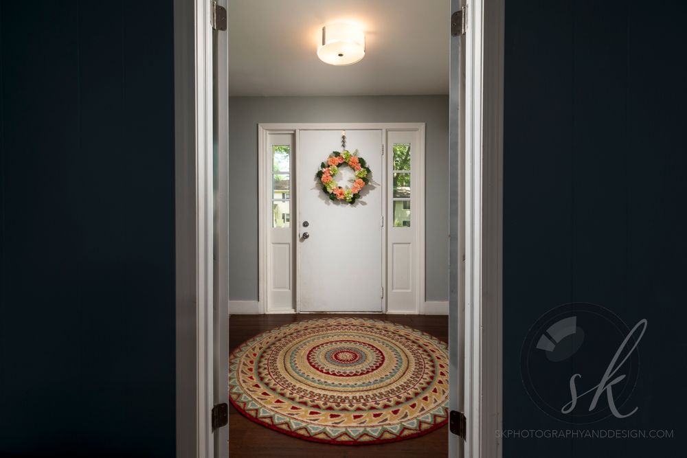 Professional Saratoga Springs real estate photo of a cozy entryway with white front door, colorful wreath, and round braided rug.