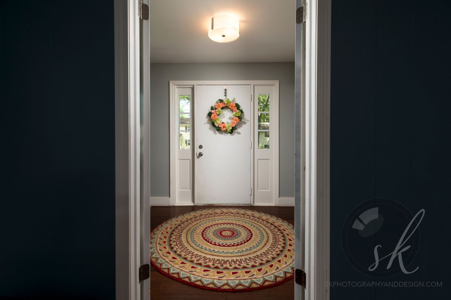 Professional Saratoga Springs real estate photo of a cozy entryway with white front door, colorful wreath, and round braided rug.