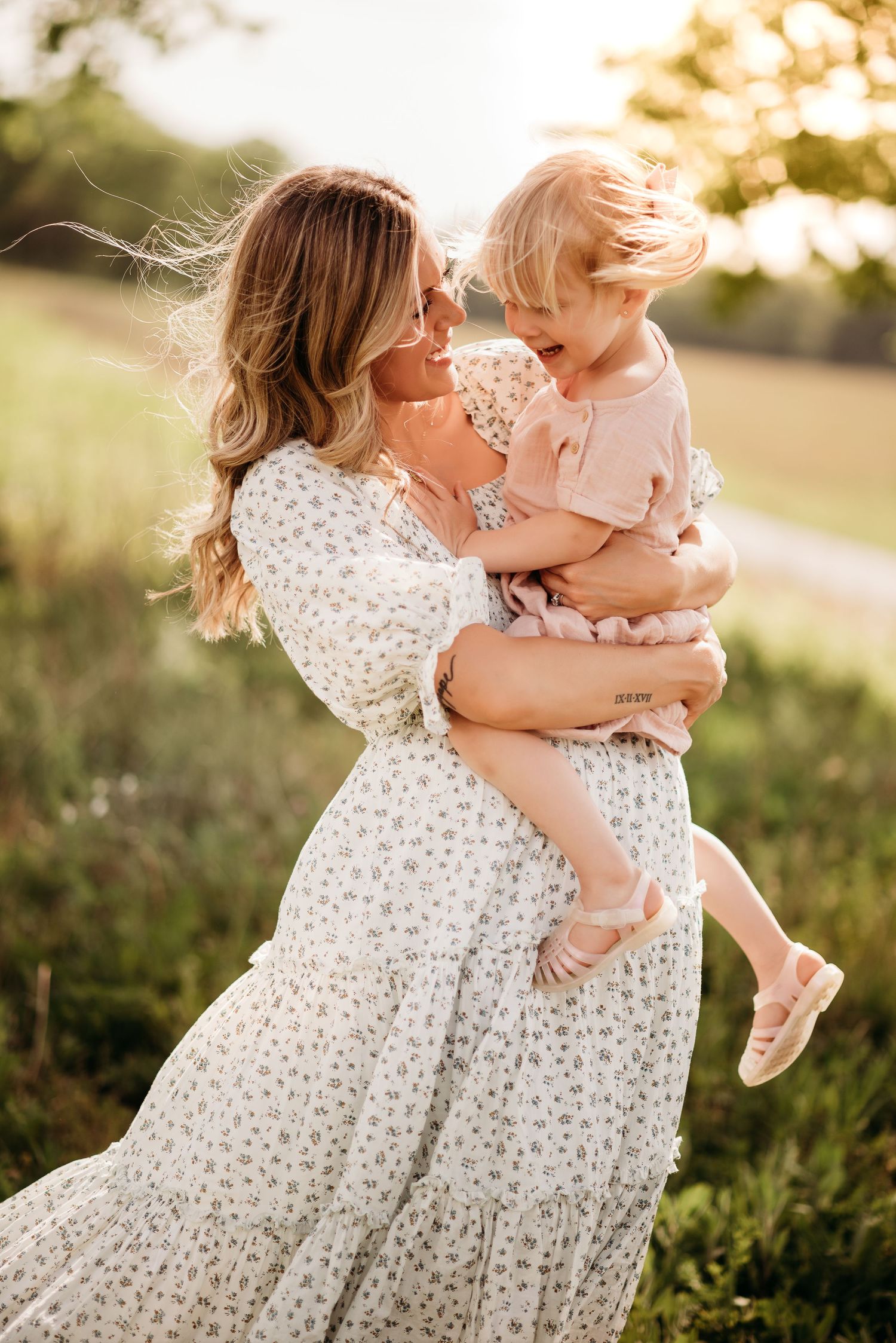 Two figures share a tender moment in a floral dress and pink outfit against golden sunlit grass at sunset.