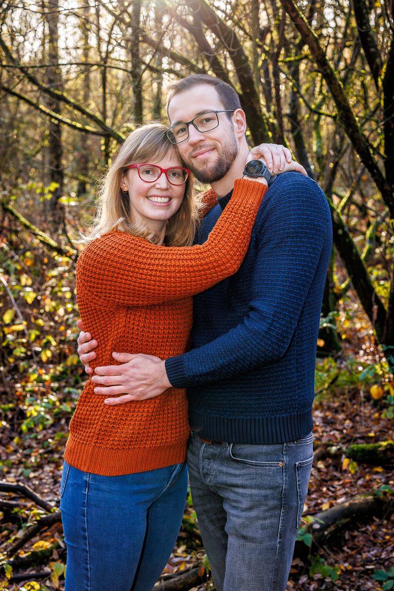 A couple in burnt orange and navy sweaters embrace lovingly among autumn trees and fallen leaves in a forest setting.