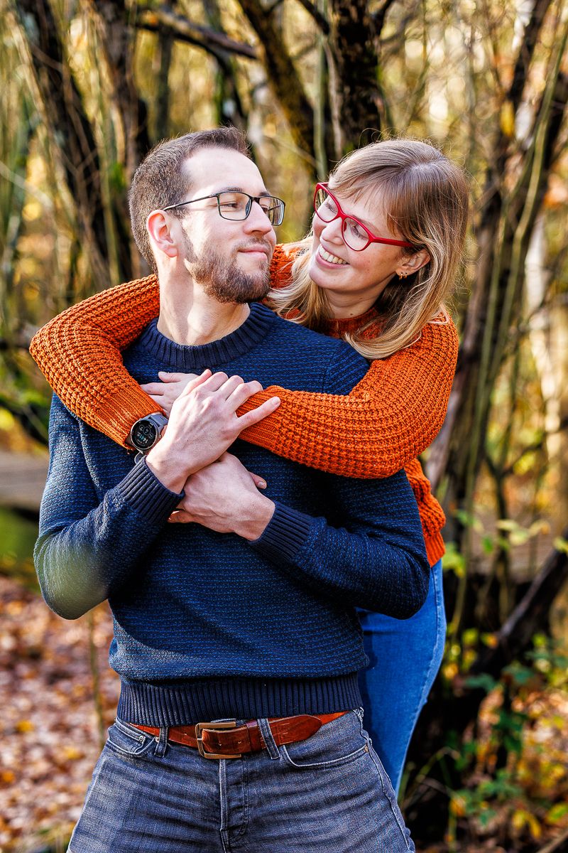 A couple embraces outdoors in autumn, wearing cozy sweaters and glasses while sharing a playful moment in the woods.