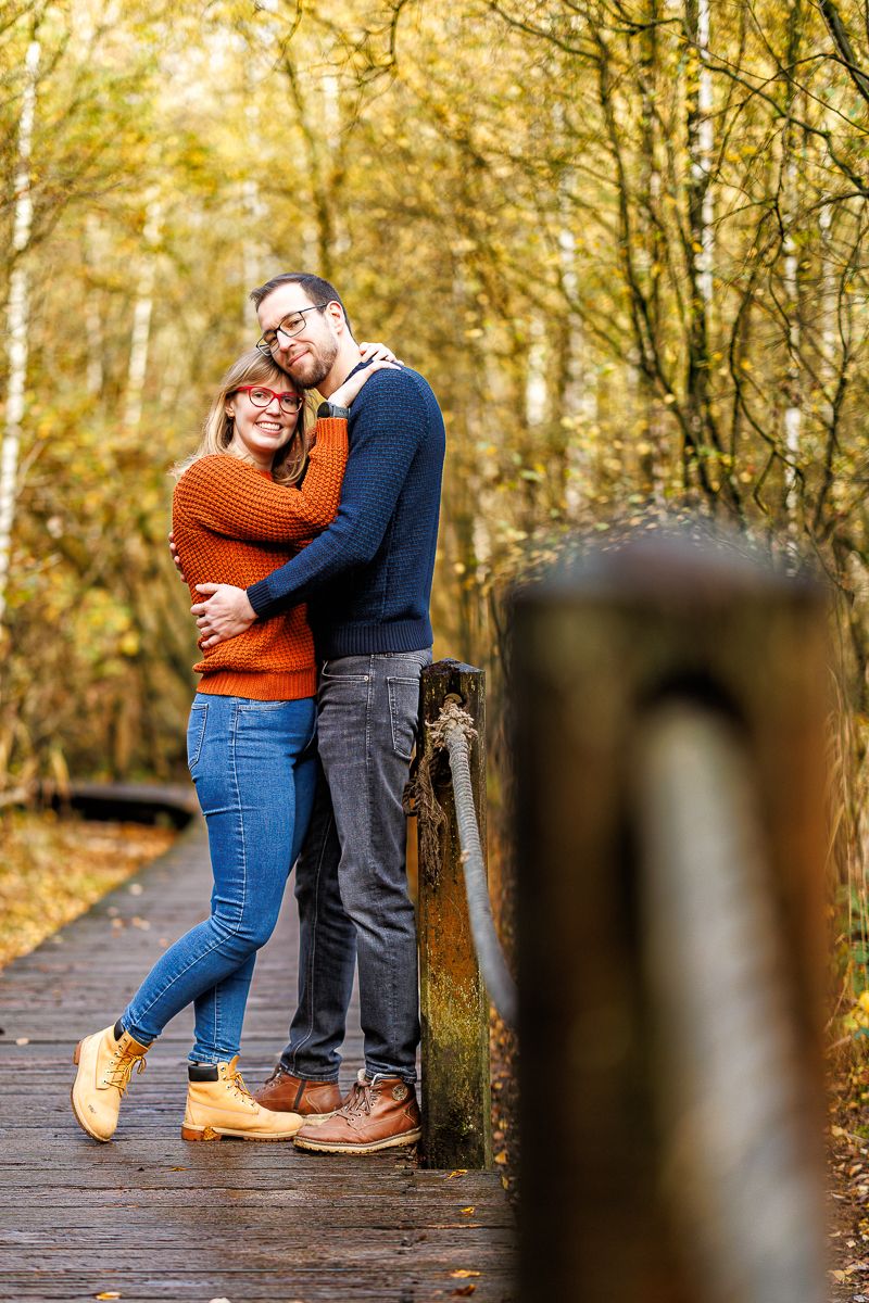 A couple embraces on a wooden boardwalk surrounded by autumn trees in a romantic outdoor setting.