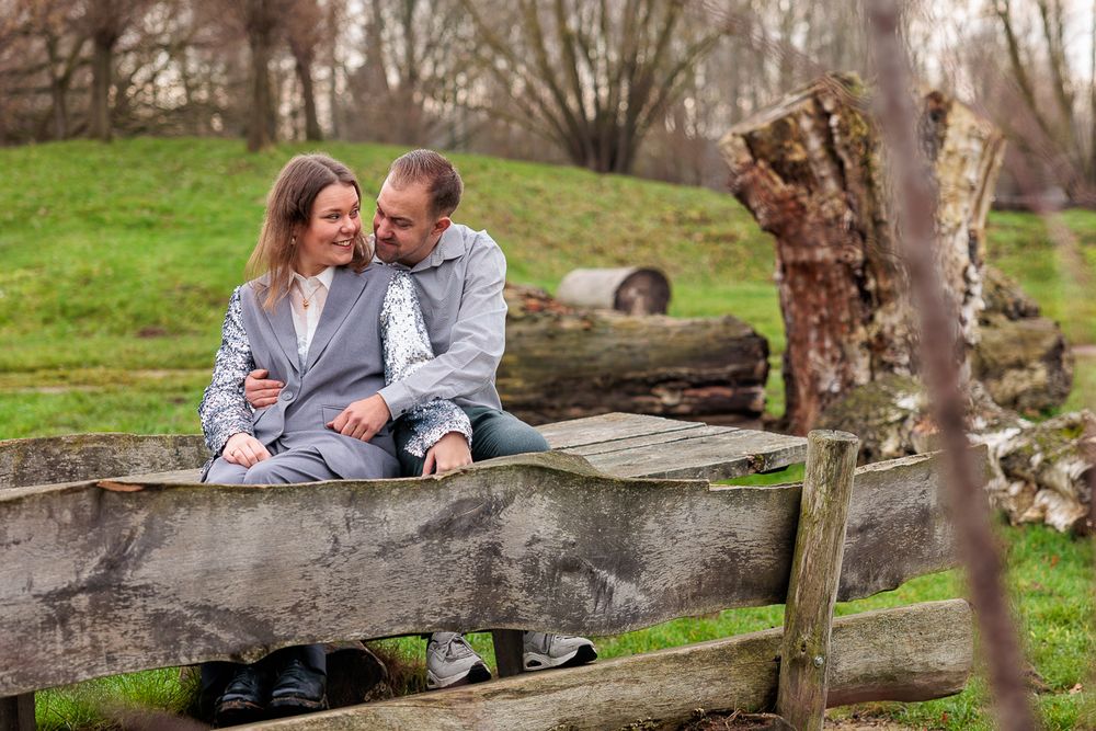 A couple sits close together on a rustic wooden bridge in a peaceful park setting during autumn.