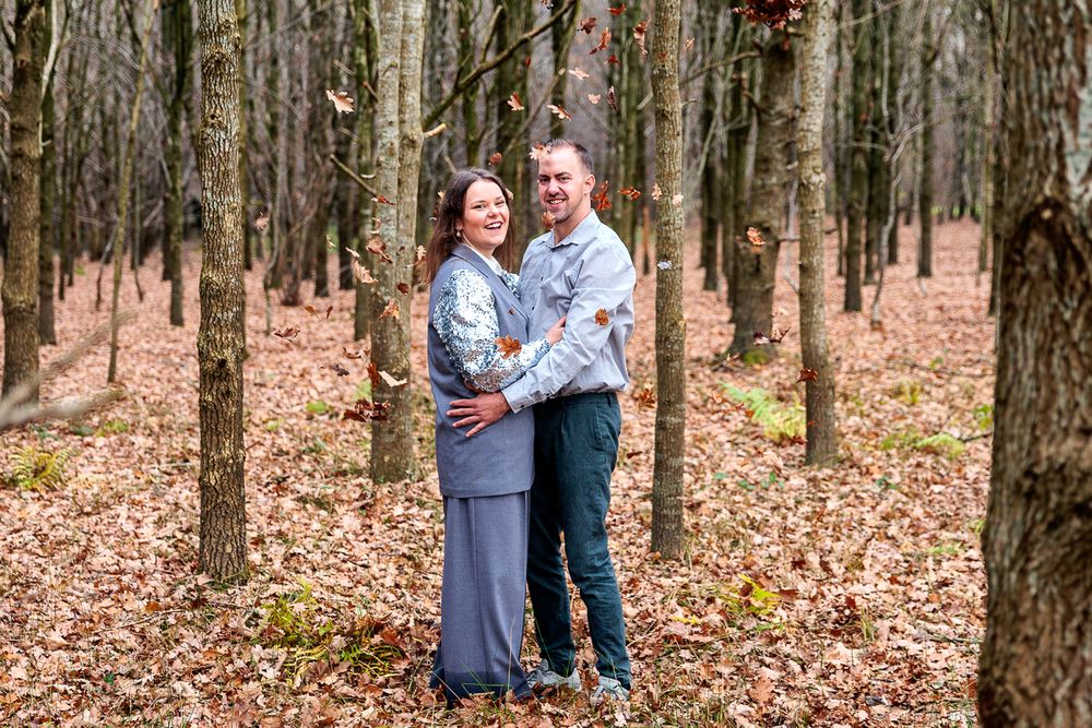 A couple shares an intimate moment while standing among tall trees in a forest covered with fallen autumn leaves.
