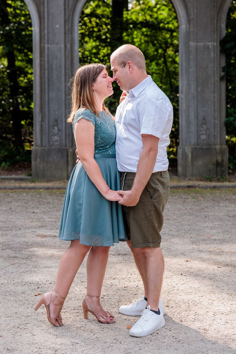 Couple in love standing face to face holding hands outdoors, woman in teal dress and man in white shirt with khaki shorts.