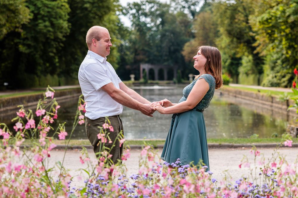 Couple holding hands facing each other in a garden with pink flowers and a pond in the background.