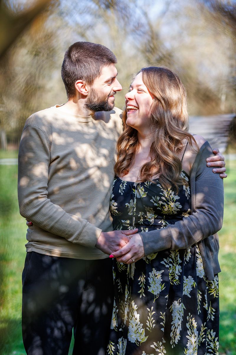 A happy couple laughing and embracing outdoors in a sunny park, man in gray sweater, woman in floral dress.