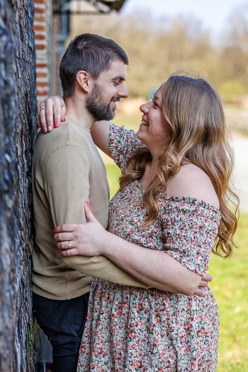 A smiling couple embraces tenderly against a brick wall outdoors, the woman in a floral dress and the man in a tan sweater.