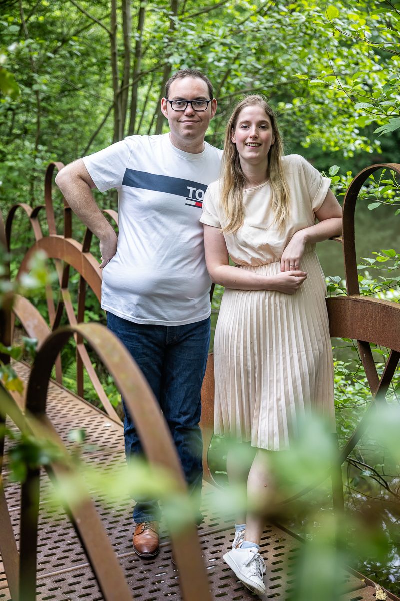 A couple poses together on a wooden footbridge surrounded by lush green foliage in a natural setting.