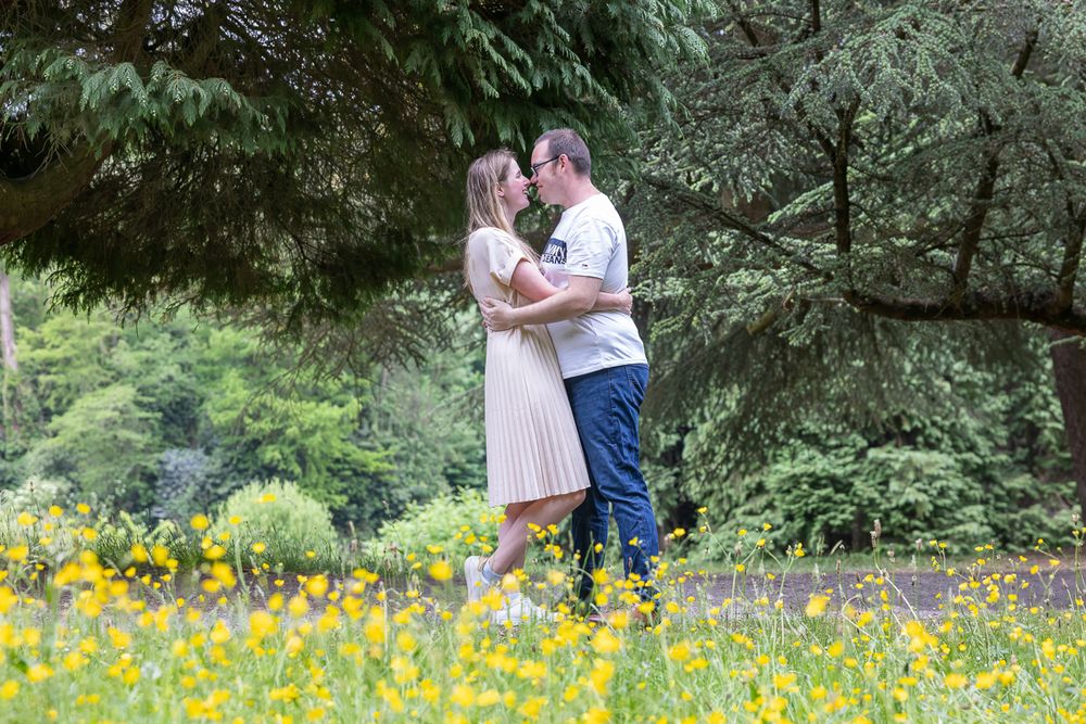 A romantic couple shares a tender moment while standing in a meadow of bright yellow wildflowers surrounded by trees.