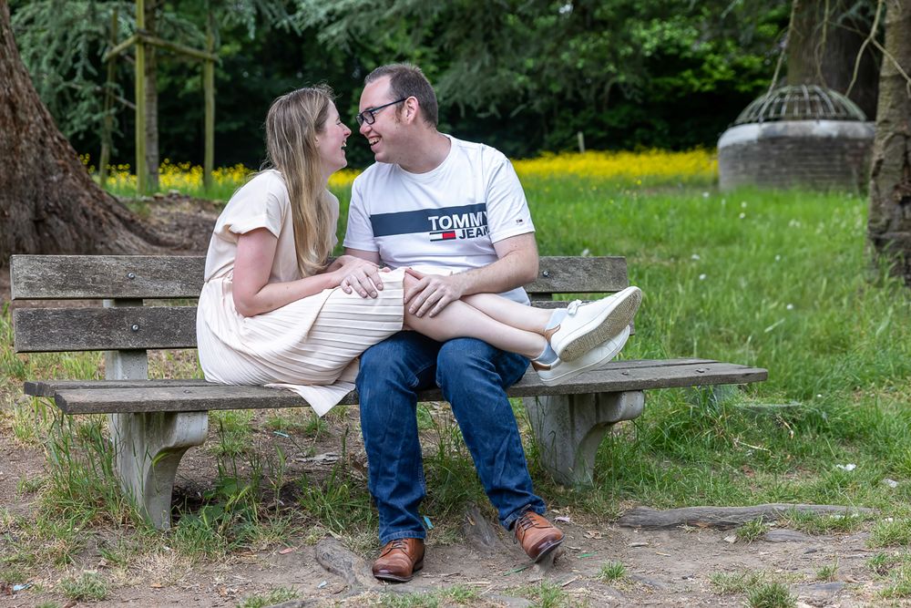 A couple shares an intimate moment while sitting together on a wooden bench in a park surrounded by yellow wildflowers.