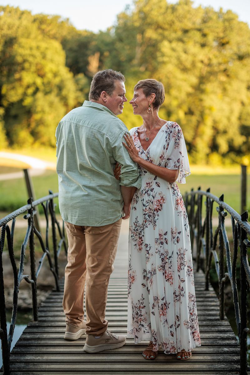 Couple smiling at each other on a wooden bridge surrounded by lush green trees in golden hour light.