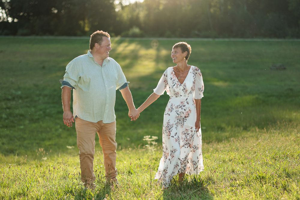 Couple holding hands walking in a sunny green field, woman in floral dress, man in light blue shirt.