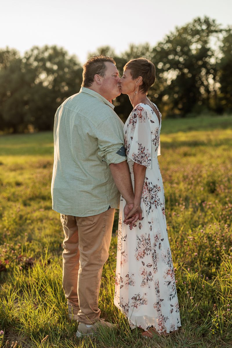 Couple sharing a romantic kiss while holding hands in a sunlit green meadow, woman in floral dress, golden hour.
