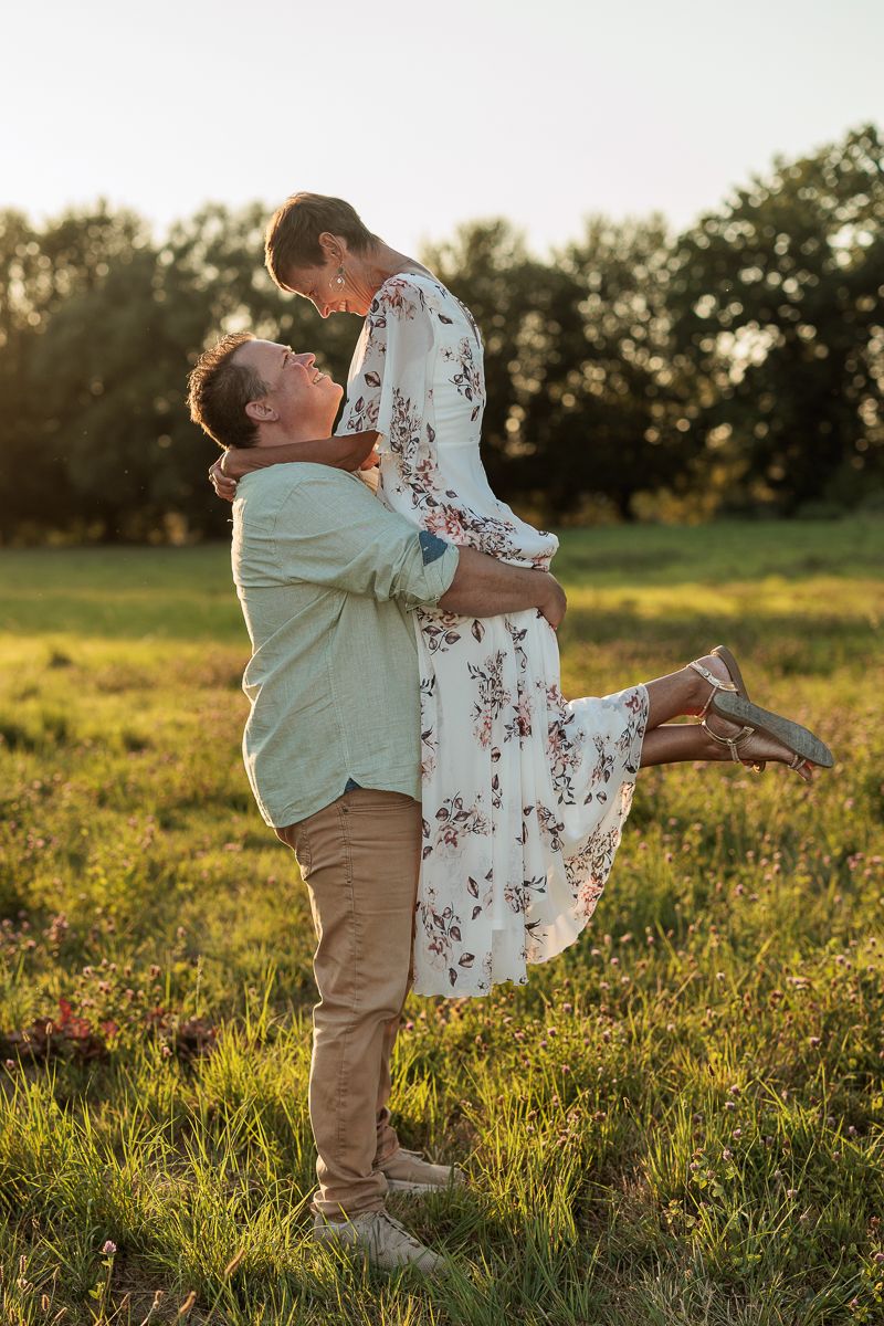 Couple sharing a romantic moment in a sunlit green field, man lifting woman in floral dress at golden hour.