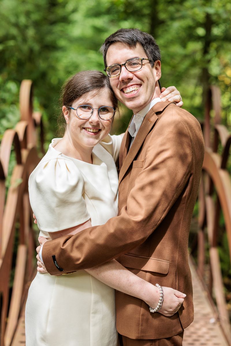 A smiling couple in wedding attire embraces on a bridge surrounded by lush greenery, wearing glasses and formal outfits.