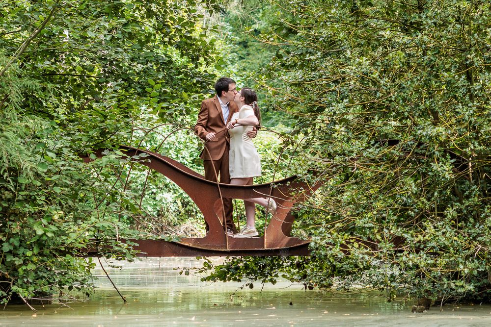 Couple sharing a romantic kiss while standing on a rustic wooden boat sculpture surrounded by lush green trees.