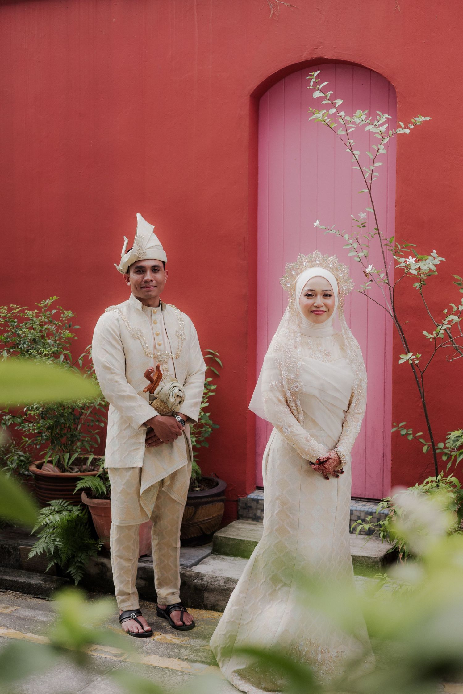 Malay couple in traditional white wedding attire standing before a vibrant red wall with a pink arched door.