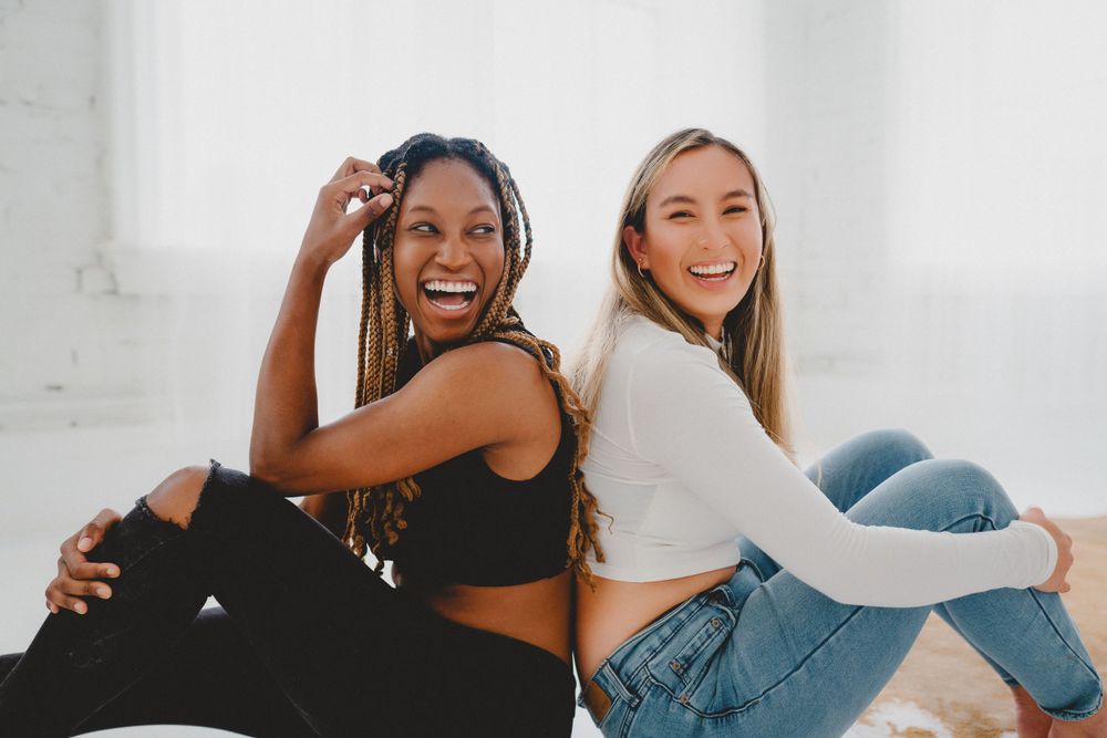 Two young women sitting back-to-back laughing joyfully in a bright white studio in Cincinnati