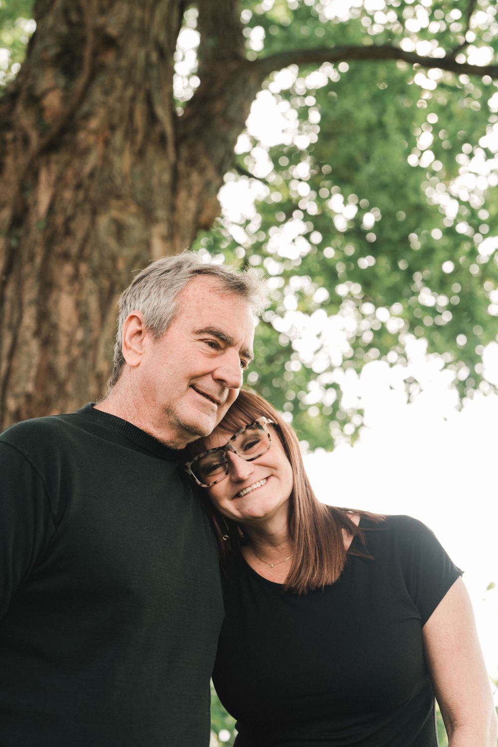 Smiling father and daughter leaning together affectionately under a large tree outdoors in Cincinnati