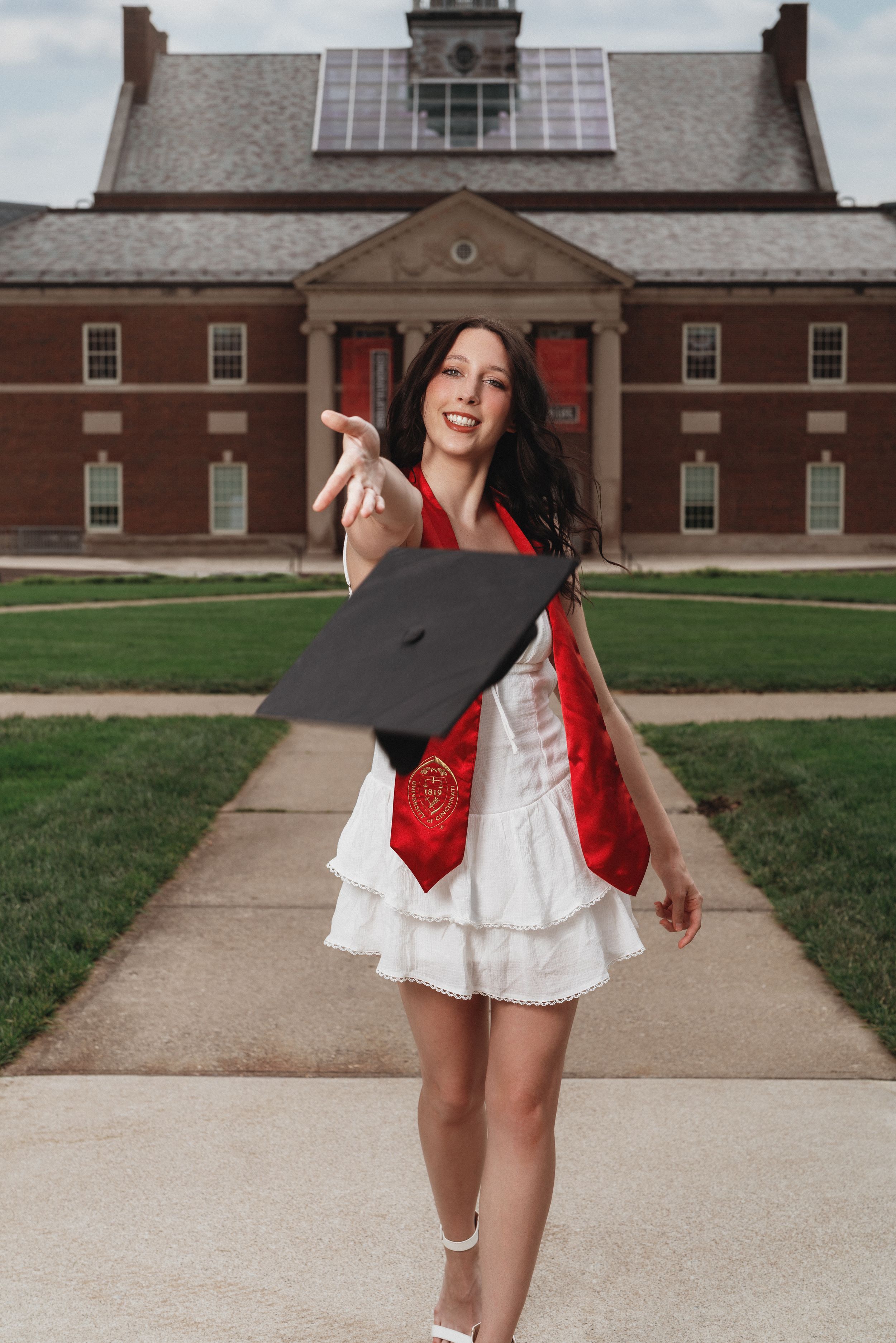 A graduate in a white dress and red sash tosses her cap in front of a brick university building in Cincinnati