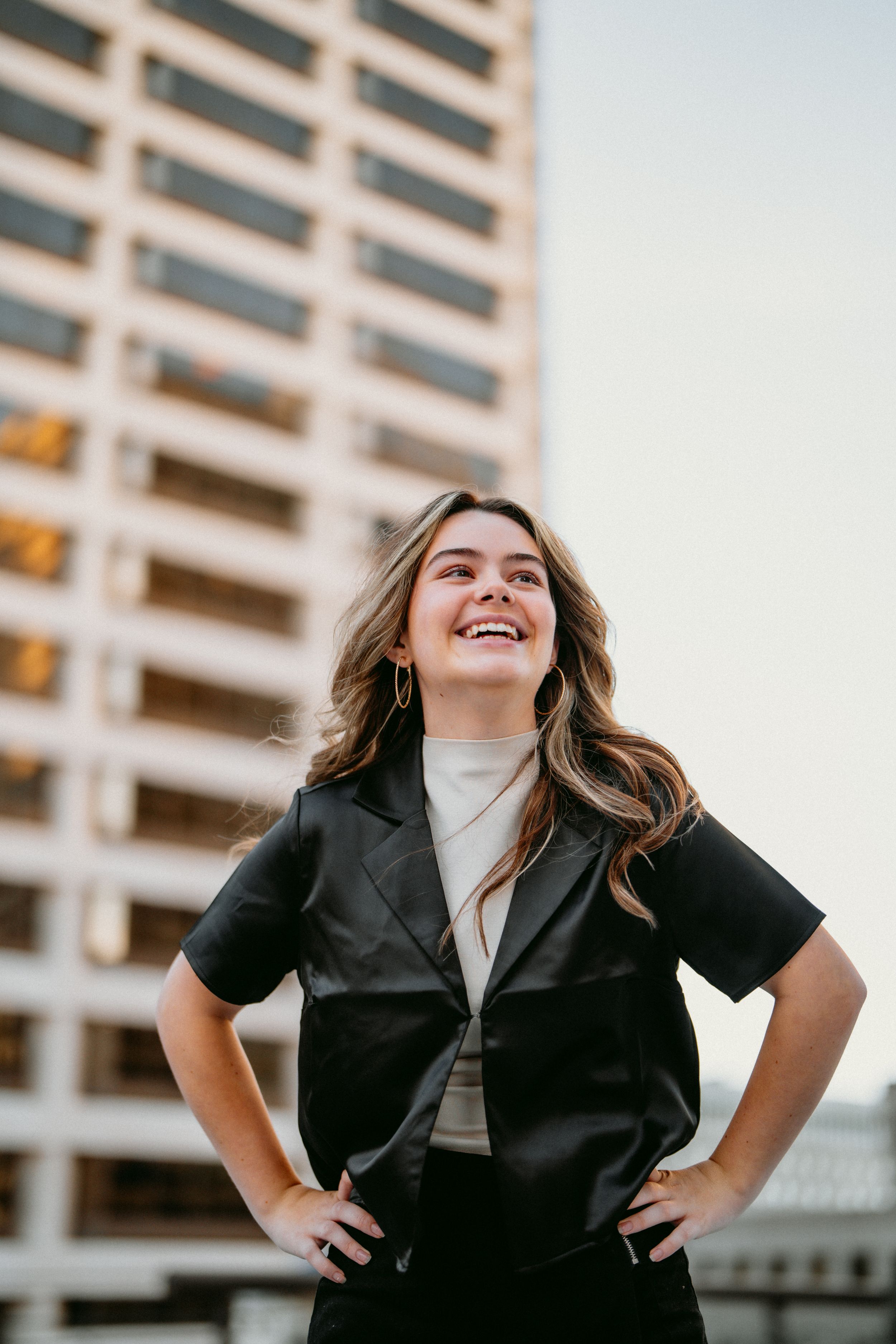 Confident young woman in black leather vest laughing with hands on hips against urban building backdrop for her senior session in Cincinnati