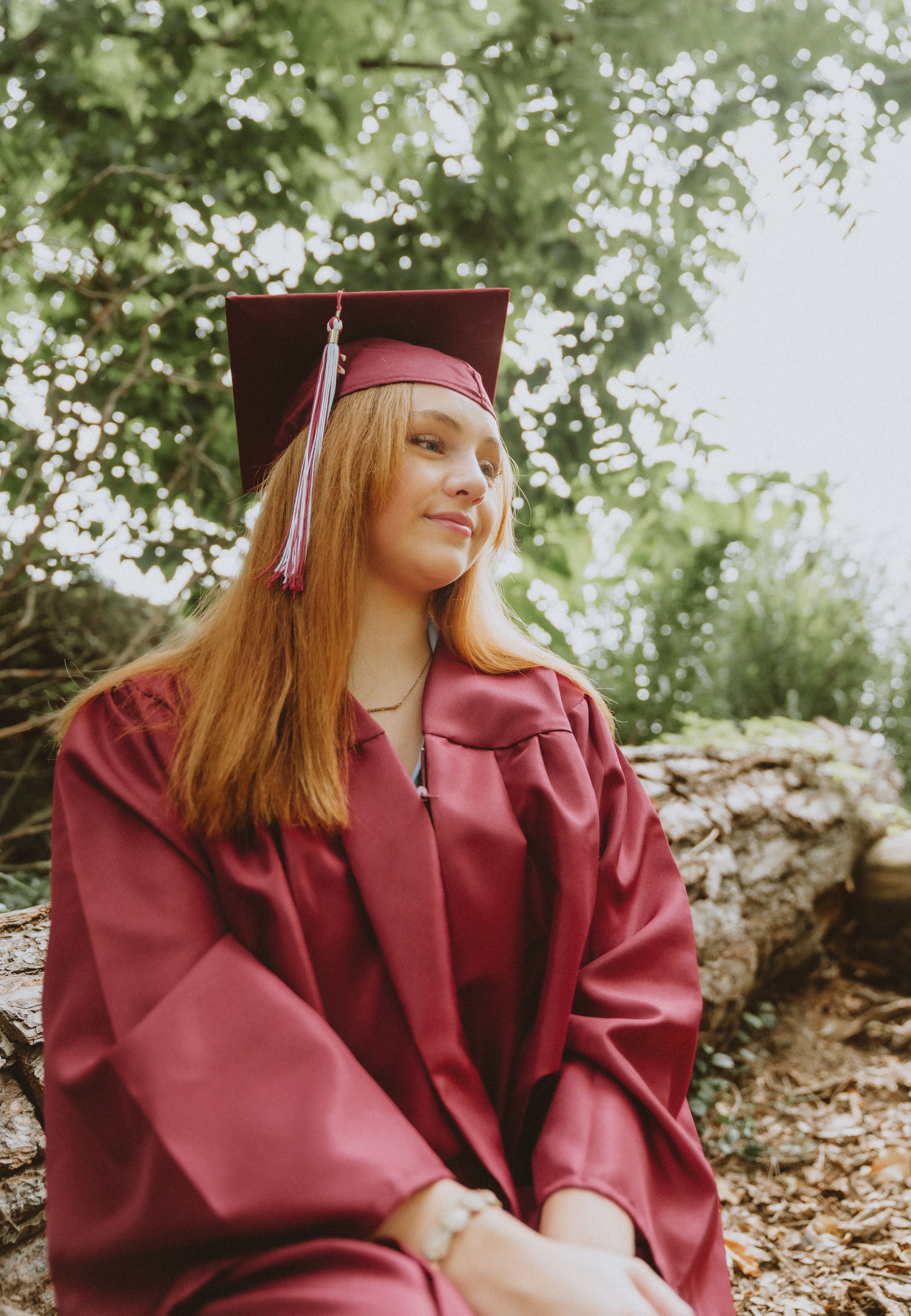 Red-haired graduate in maroon cap and gown sitting outdoors among green trees for graduation portrait in Cincinnati