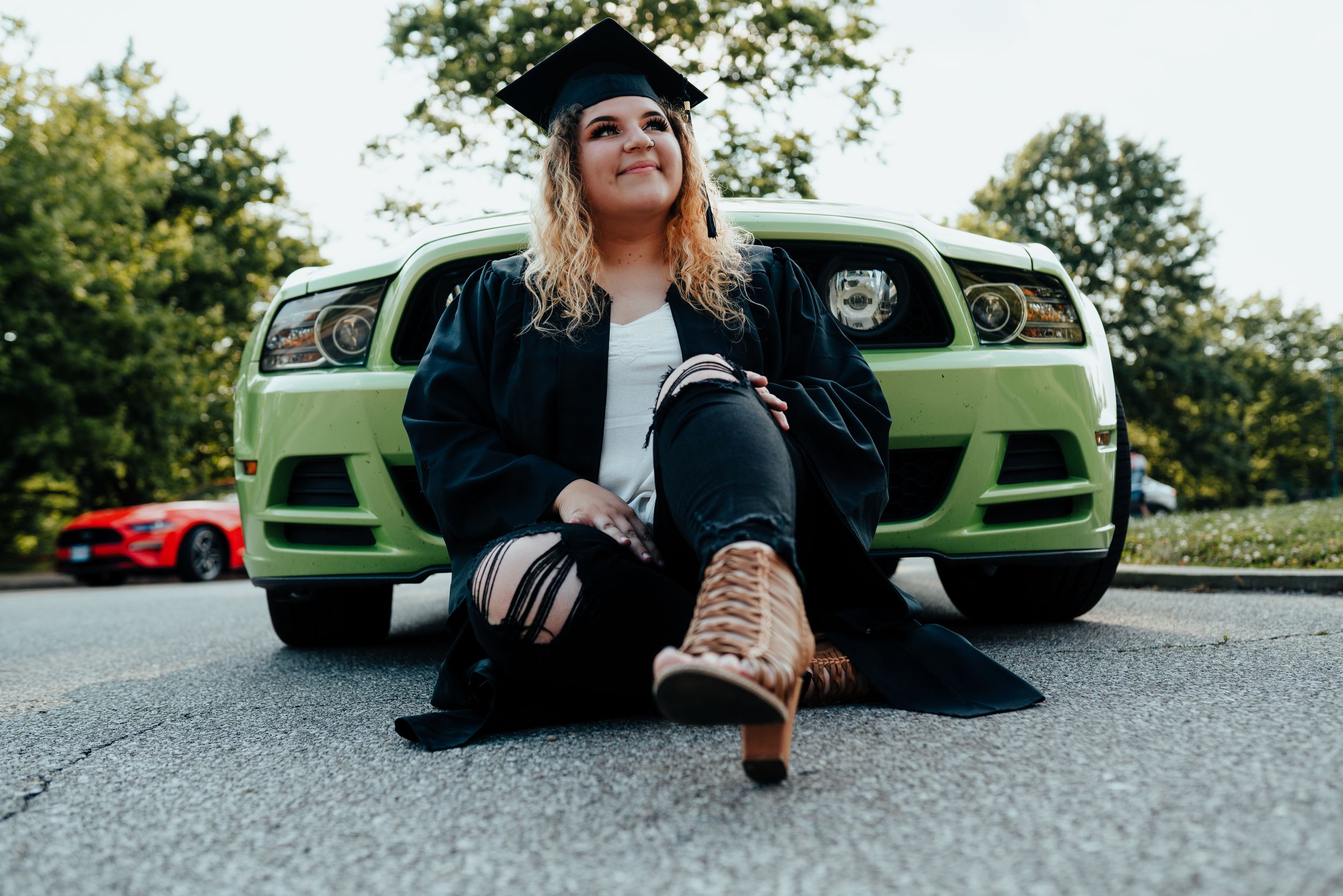 Female graduate in cap and gown sitting in front of green Mustang car for graduation photos for her senior session in Cincinnati